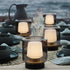Trådlös Ambience bordslampa från sackit med varm belysning i mörk glasram på dukat bord vid havet tillsammans med flera tända lampor