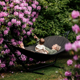 Svart utomhushängmatta med stativ där en vuxen och ett barn kopplar av i en blomstrande trädgård med rosa blommor.