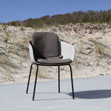 Mörkgrå sittdyna i en modern uteplatsstol med vita armstöd och svarta ben framför sanddyner och strandgräs i nordiskt solljus