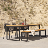 sackit utomhusmatgrupp med svarta stolar, bord och bänk i trä och metall, placerad framför sanddyner med strandgräs och en filt över bänken