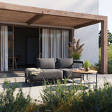 Mörkgrå modulär loungesoffa med öppen avslutning åt vänster på en solig uteplats under en pergola designad av sackit för nordiskt uteliv