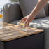 Teakbord med förvaring intill en grå utomhussoffa medan en hand ställer ner ett glas citronvatten på bordet från sackit