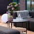 Runt svart utomhussoffbord från sackit med bordsskiva i pulverlackerad aluminium och hylla i teak framför en loungesoffa på en grönskande terrass