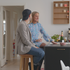 En person sitter på en hög barstol i ek vid en köksö tillsammans med en annan person i ett ljust nordiskt hem