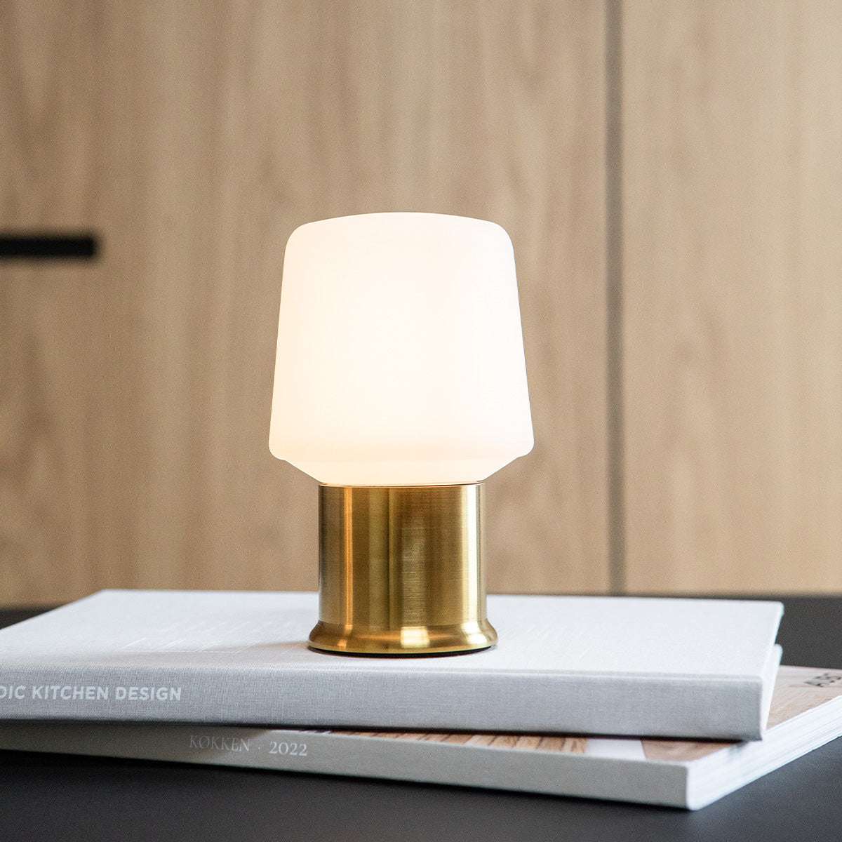 Ambience lampfot i mässingsfärg med vit lampskärm på staplade böcker i varm inomhusmiljö från sackit