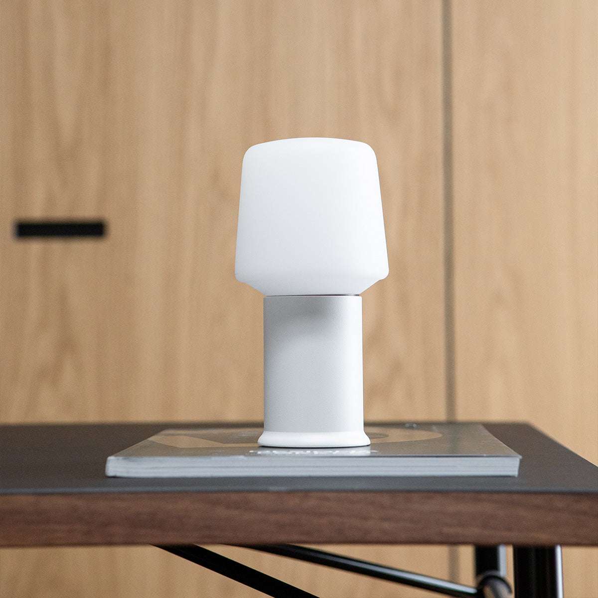 Vit bordslampa med slimmad lampfot i rostfritt stål på ett mörkt träbord från sackit