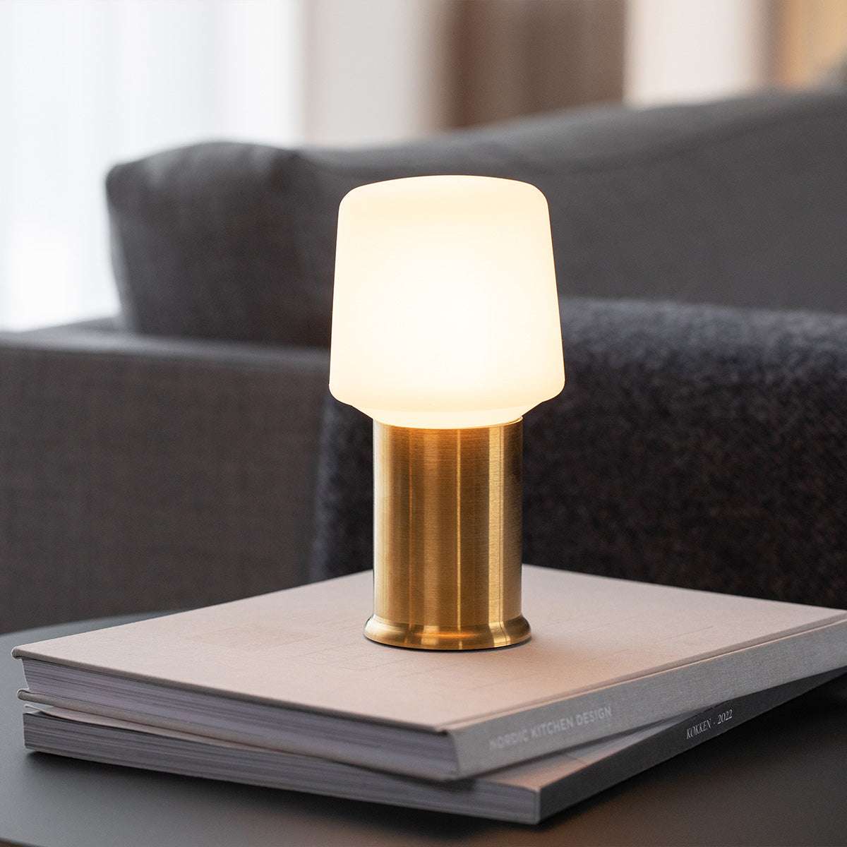 sackit Ambience bordslampa med guldfärgad metallfot och varmt lysande vit lampskärm på staplade böcker i ett vardagsrum