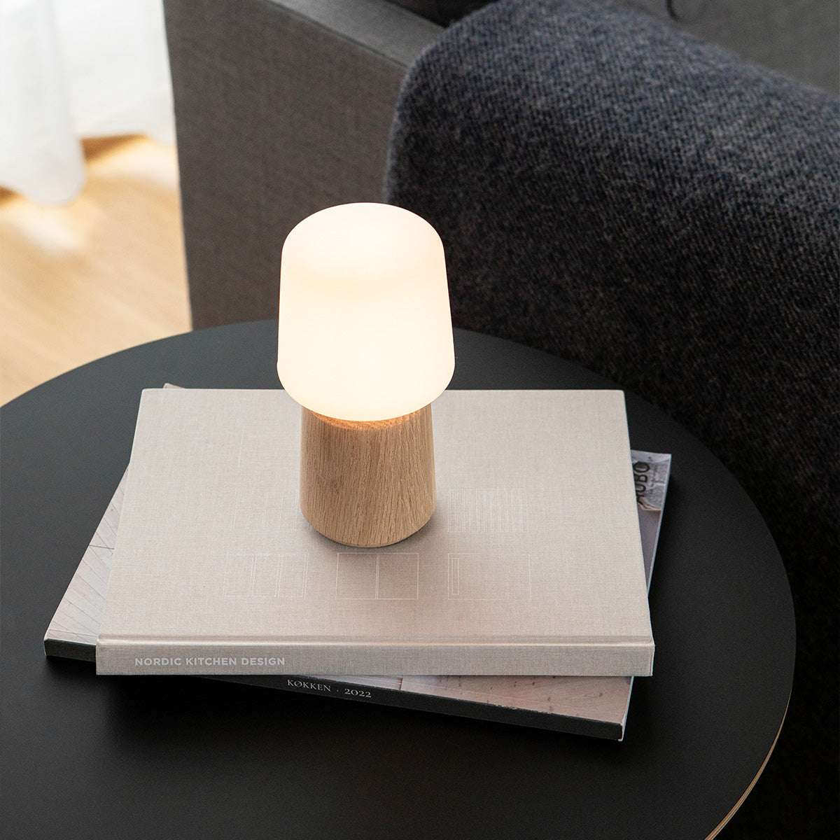 Bordslampa med ljus ekfot och varmvit skärm på ett svart runt bord i vardagsrumsmiljö från sackit