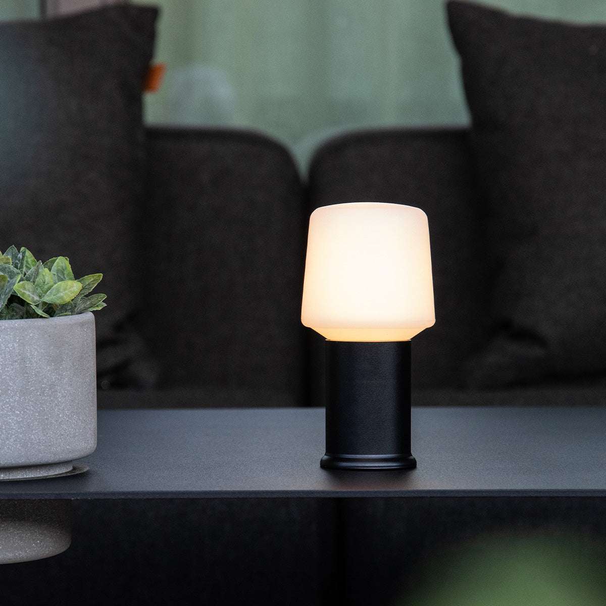 Glödande trådlös bordslampa från sackit med vit lampskärm och svart bas på ett mörkt bord framför svarta kuddar och en grön växt