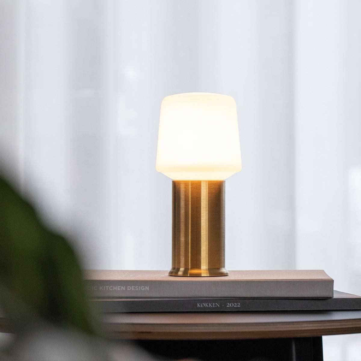 Trådlös bordslampa från sackit med varmt vitt sken och cylindrisk metallfot på staplade böcker i en ljus inomhusmiljö