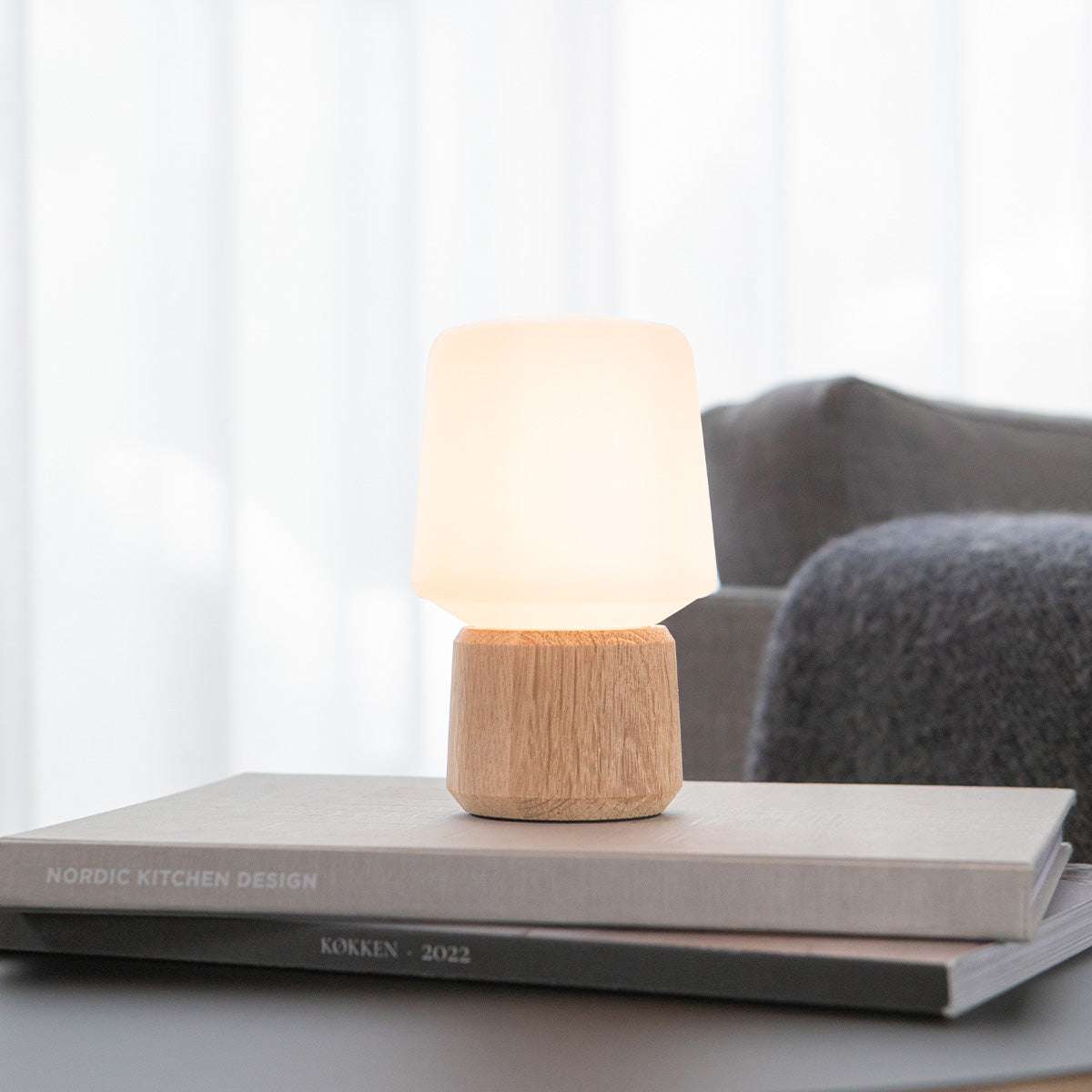Trådlös bordslampa från sackit med varmvit kupa och träfärgad fot placerad på staplade böcker i ett ljust vardagsrum