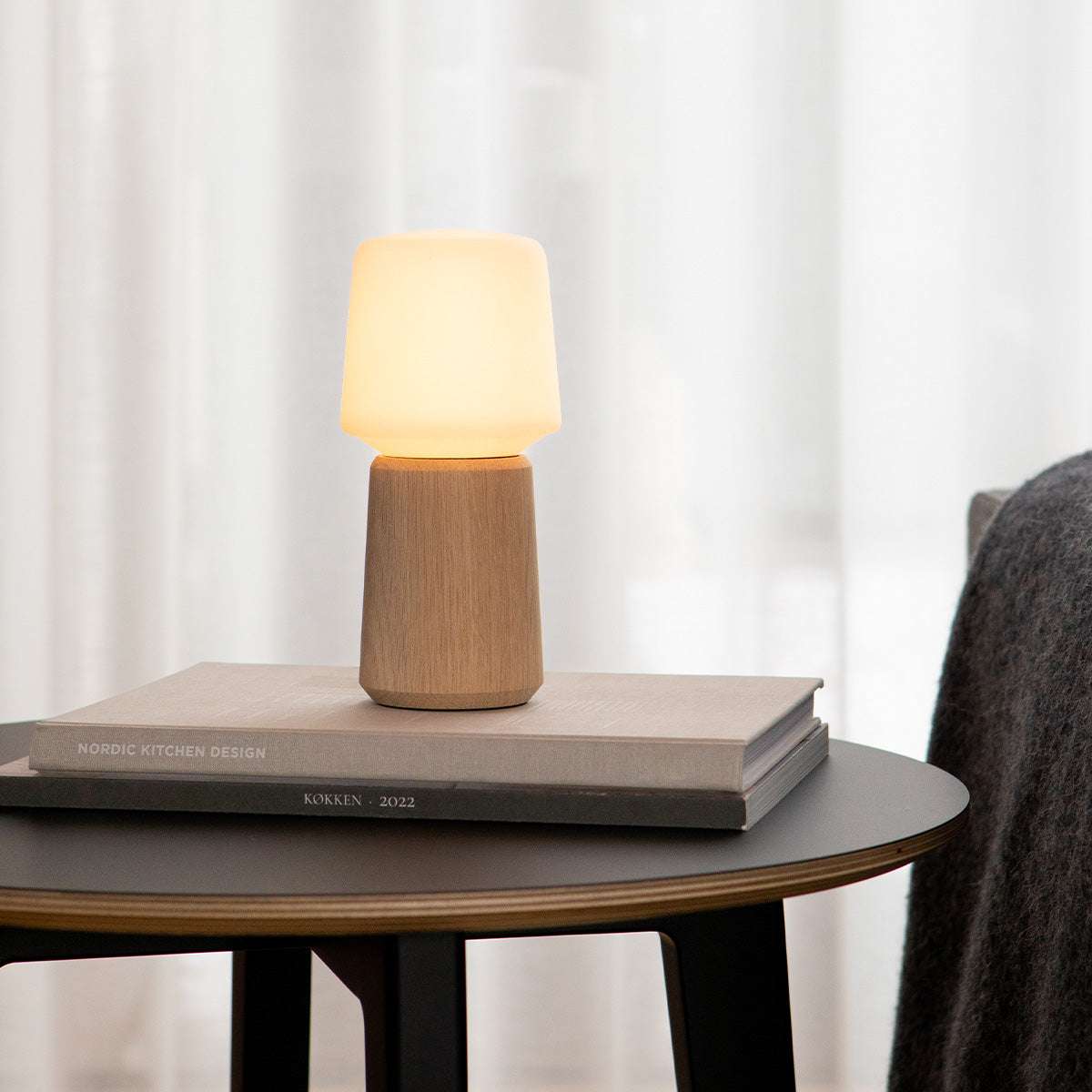 Trådlös bordslampa från sackit med varm belysning och träfärgad bas på ett runt sidobord i nordisk inredning framför ljusa gardiner