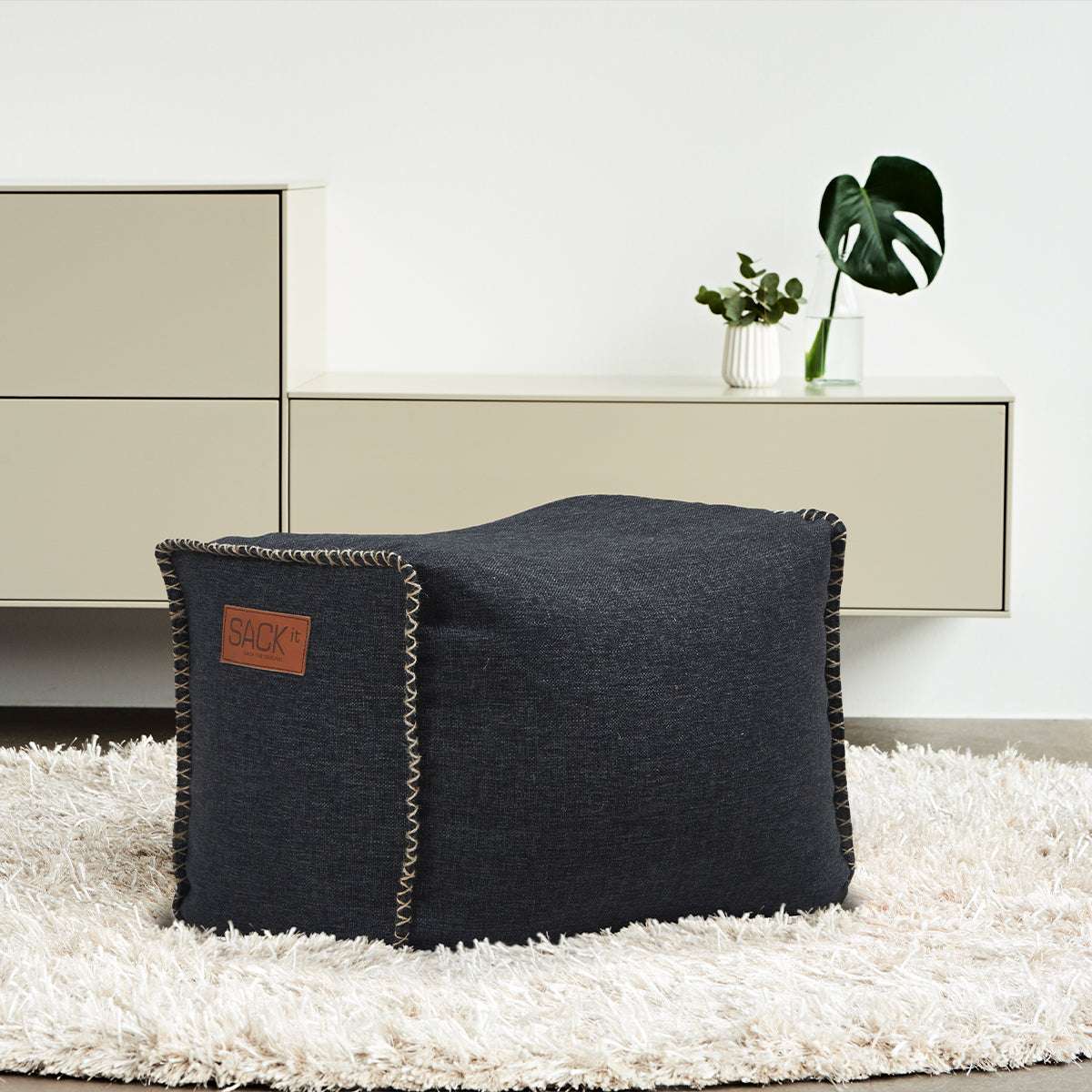 Mörkgrå fyrkantig sittpuff från sackit i ett ljust vardagsrum på en mjuk ryamatta med stilren förvaring och gröna växter i bakgrunden
