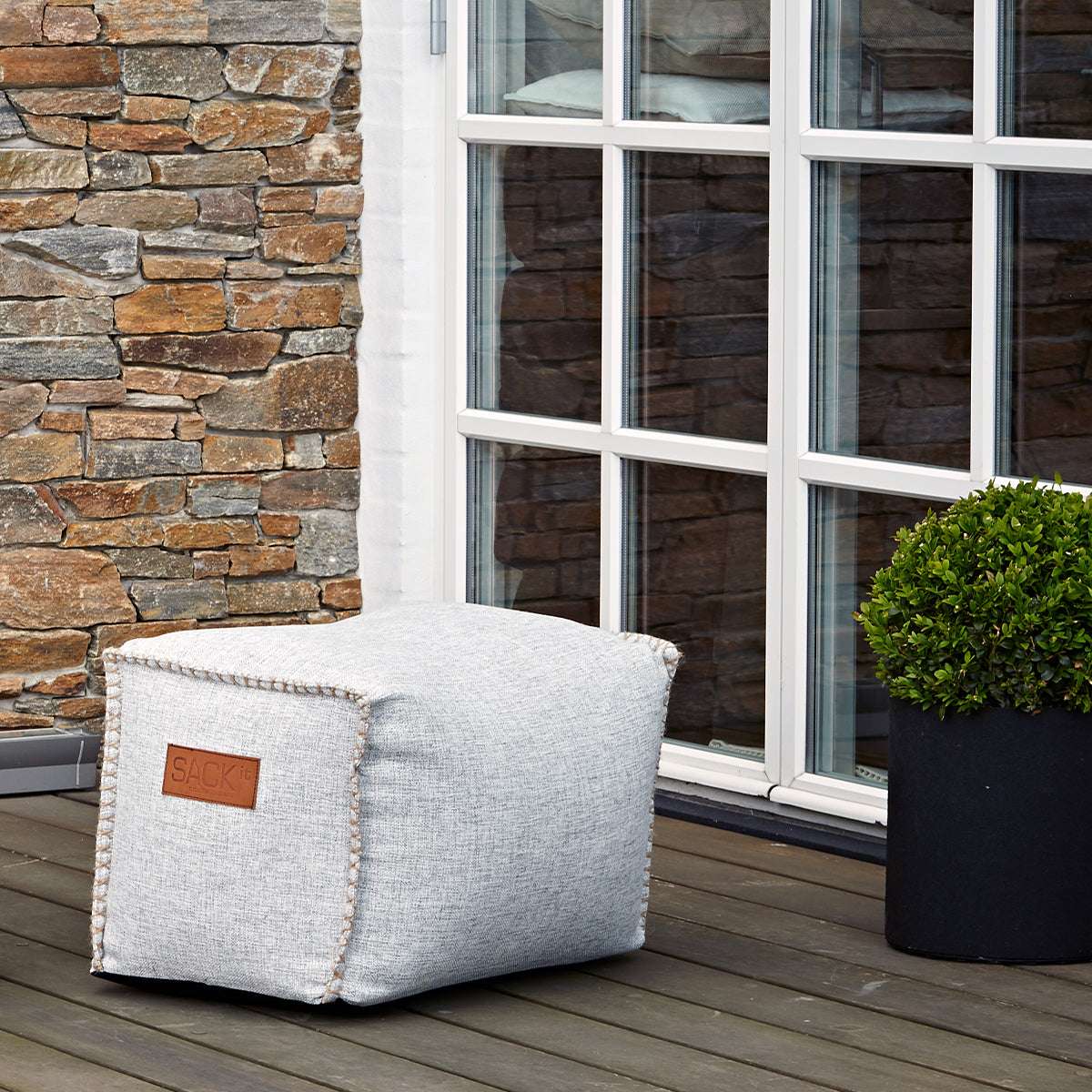 Ljusgrå fyrkantig sittpuff från sackit på en träaltan framför ett fönster och en stenmur med en grön växt i kruka intill en modern nordisk uteplats