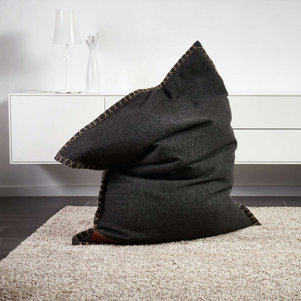 Mörkgrå Cobana beanbag i slitstarkt tyg med dekorativa sömmar står som en bekväm loungestol på en ljus matta i ett modernt vardagsrum från sackit