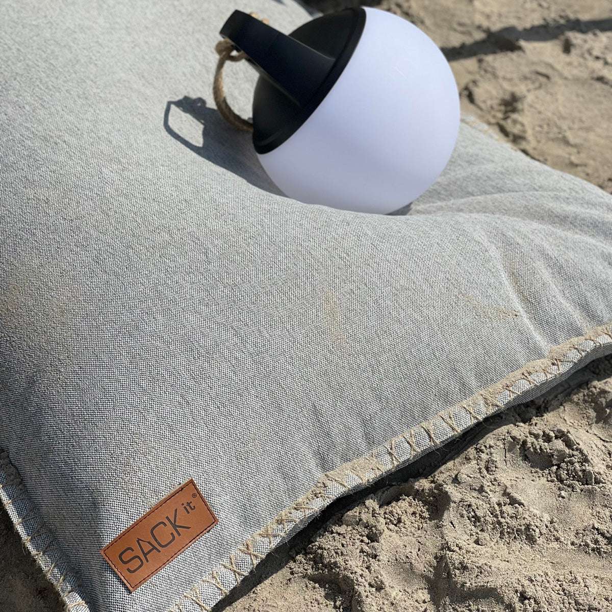 Grå sittsäck från sackit på en sandstrand med en vit rund lampa ovanpå. Slitstarkt tyg och fransad kant för avkoppling utomhus.