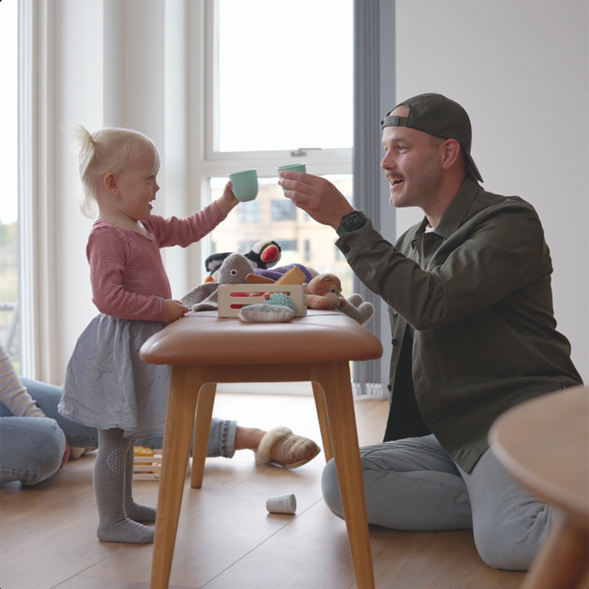 Barn och vuxen leker tebjudning vid ett ljust träbord i ett nordiskt hem med leksaker och mjuka färger för en varm och lekfull stund