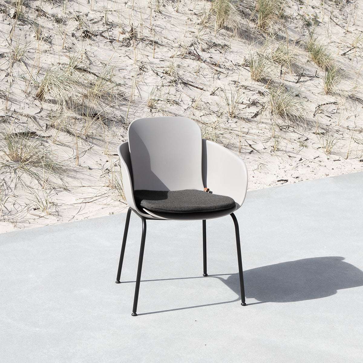 Grå utomhusstol med svart sittdyna på en ljus terrass framför sanddyner och strandgräs. Bekväm design för nordiskt väder från sackit