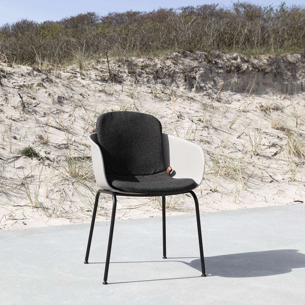 Svart sittdyna i en modern utefåtölj på en ljus terrass framför sanddyner och strandgräs