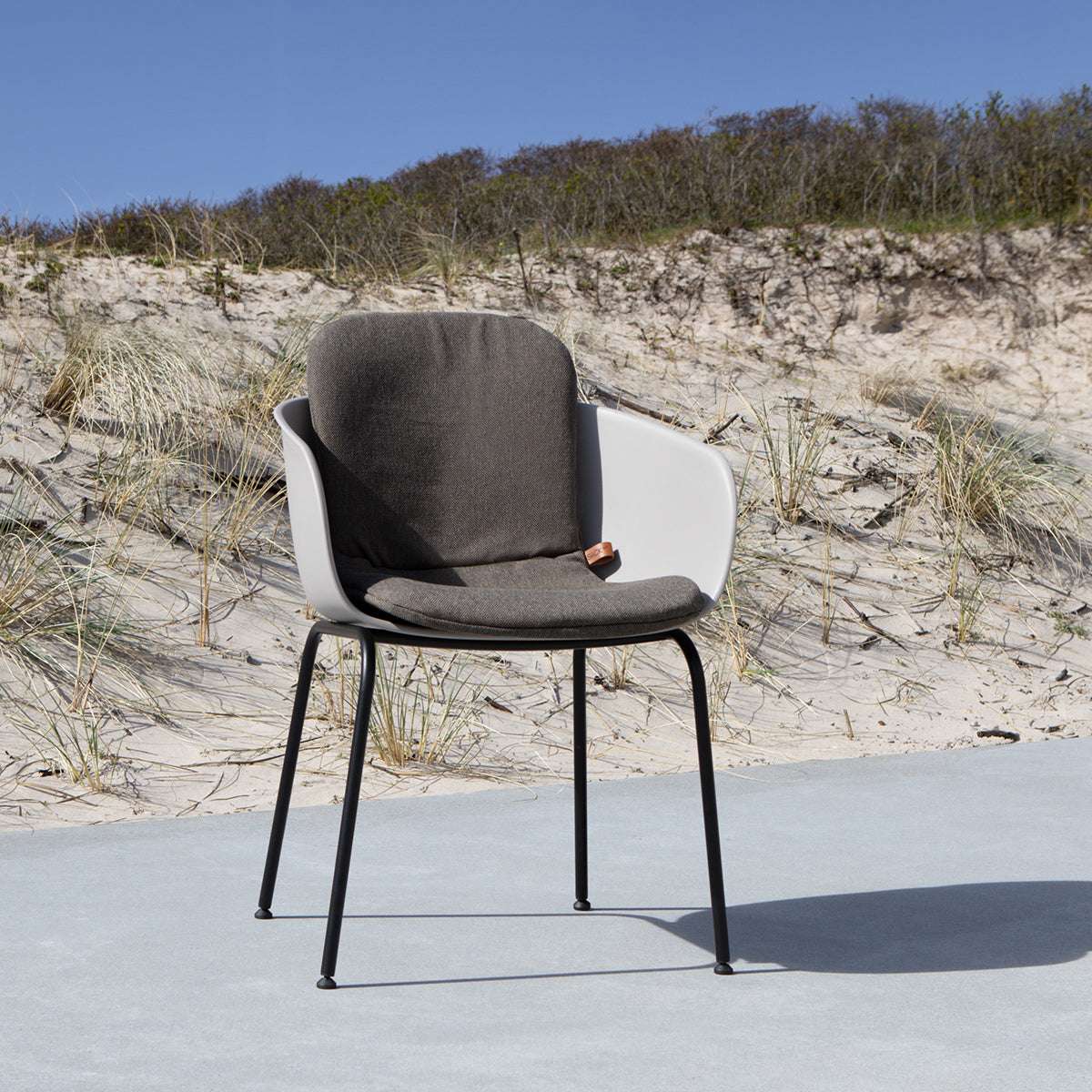 Mörkgrå sittdyna i en modern uteplatsstol med vita armstöd och svarta ben framför sanddyner och strandgräs i nordiskt solljus